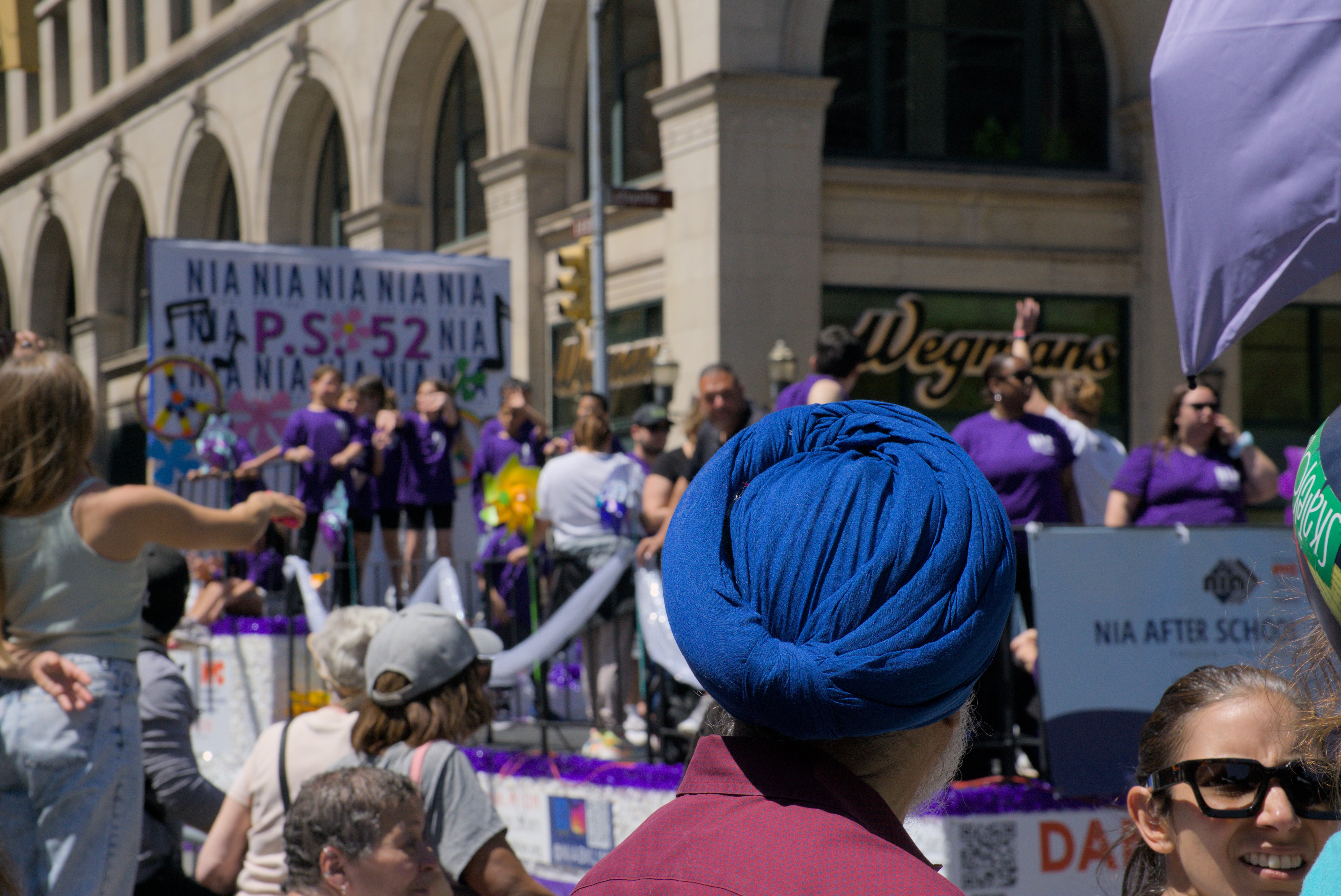 A man in a royal-blue dastar, seen from behind, watches a float of children in purple shirts on a sunny Manhattan street.